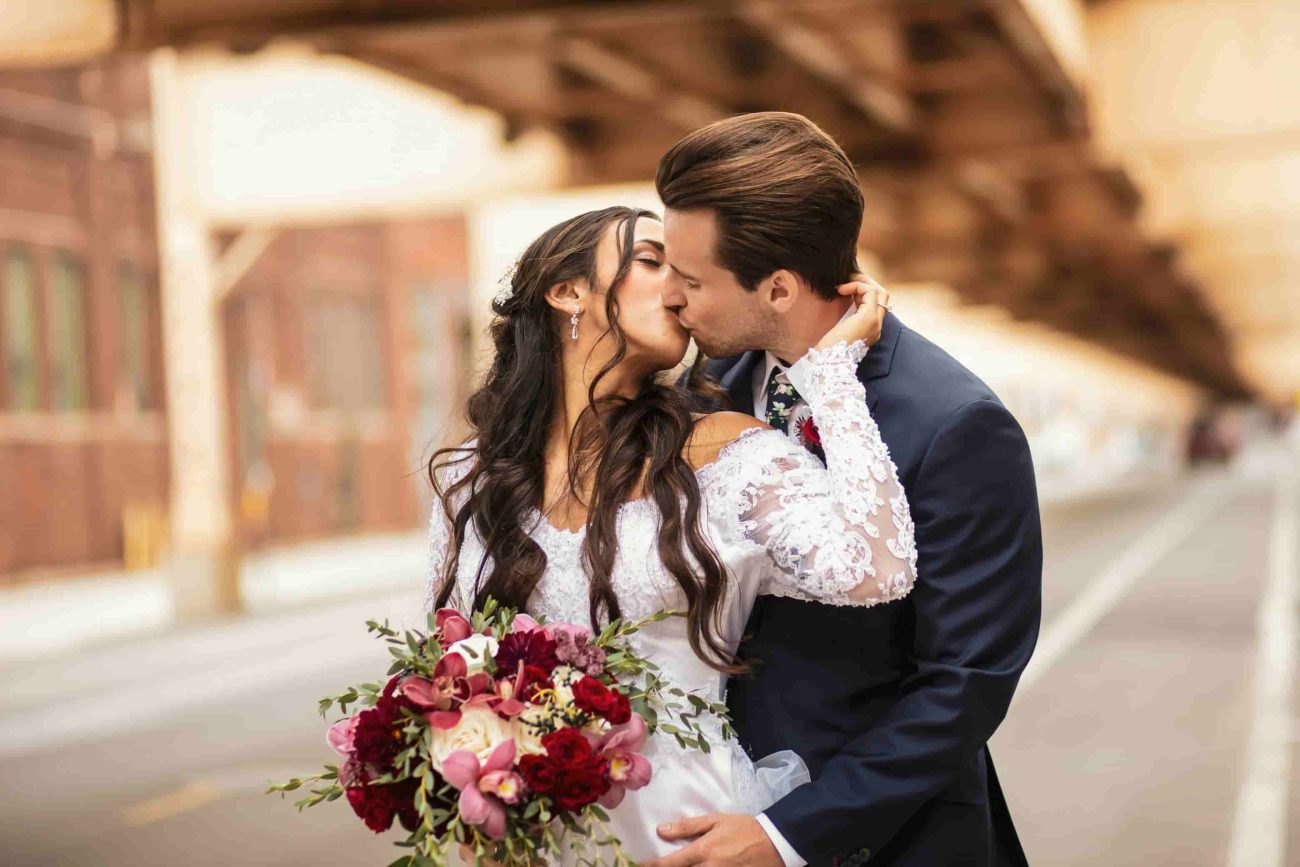 A imagem mostra um casal de noivos se beijando com um buquê de flores coloridas.