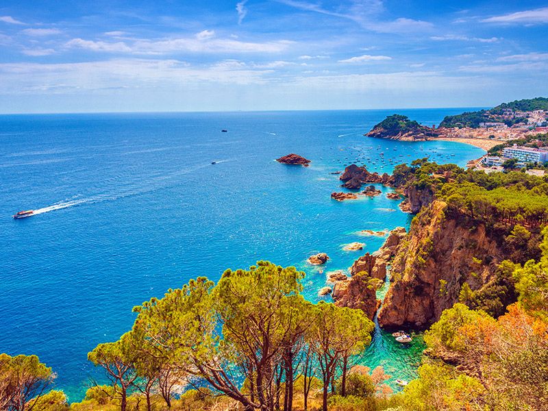 Praia na Espanha, de águas azuis, árvores ao redor e barcos navegando.