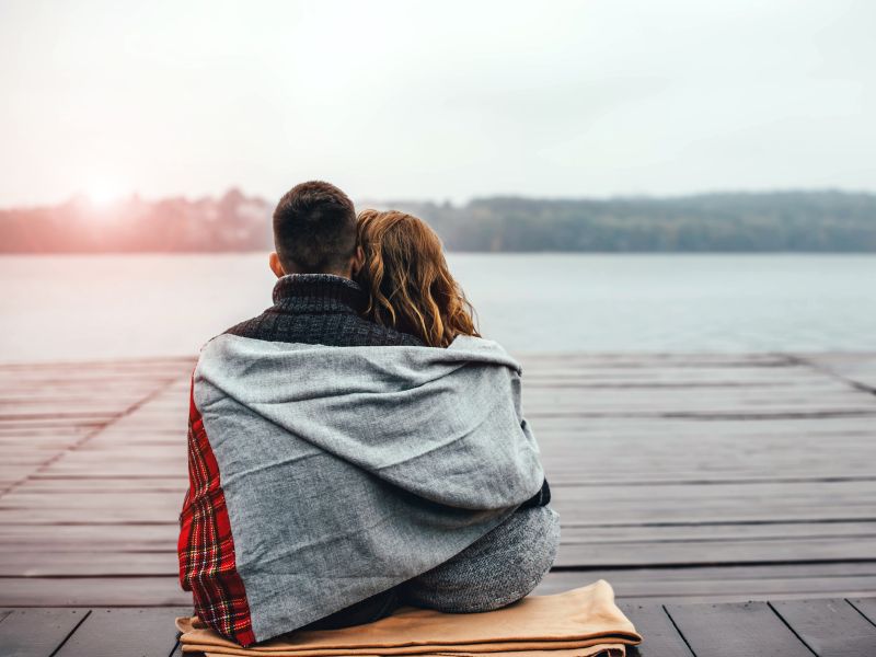 Bodas de algodão - Um casal de homem e mulher olhando um lago, sentados lado a lado, abraçados e enrolados em um cobertor