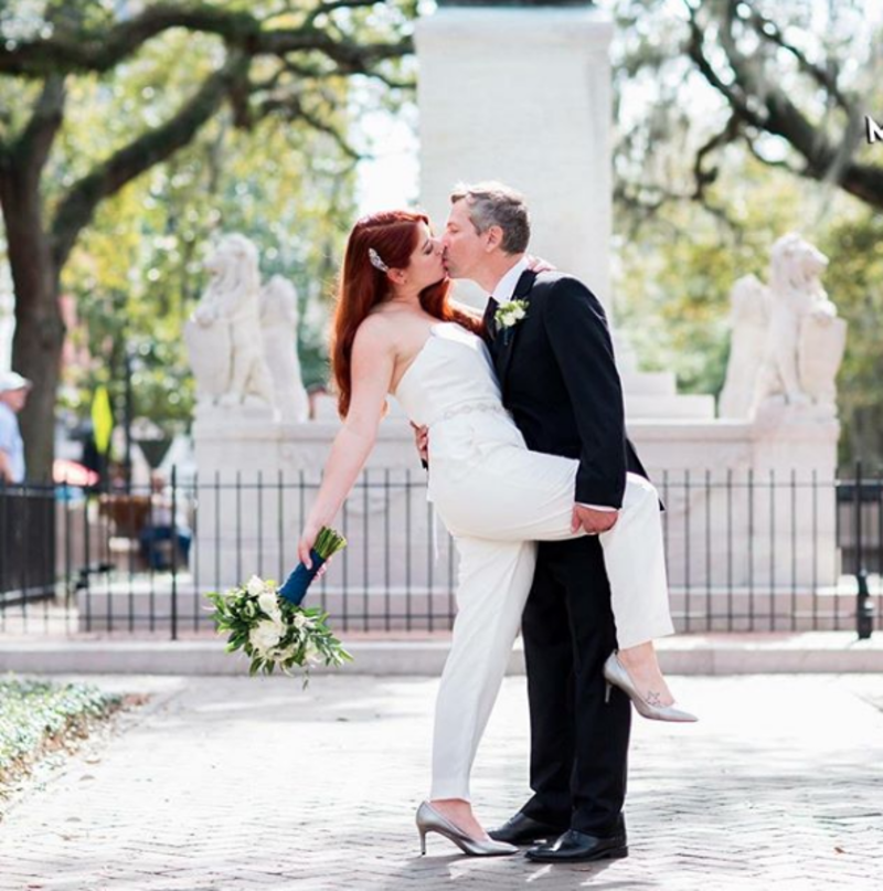 Casal se beijando na frente de uma fonte. A noiva veste macacão e segura o buquê. 