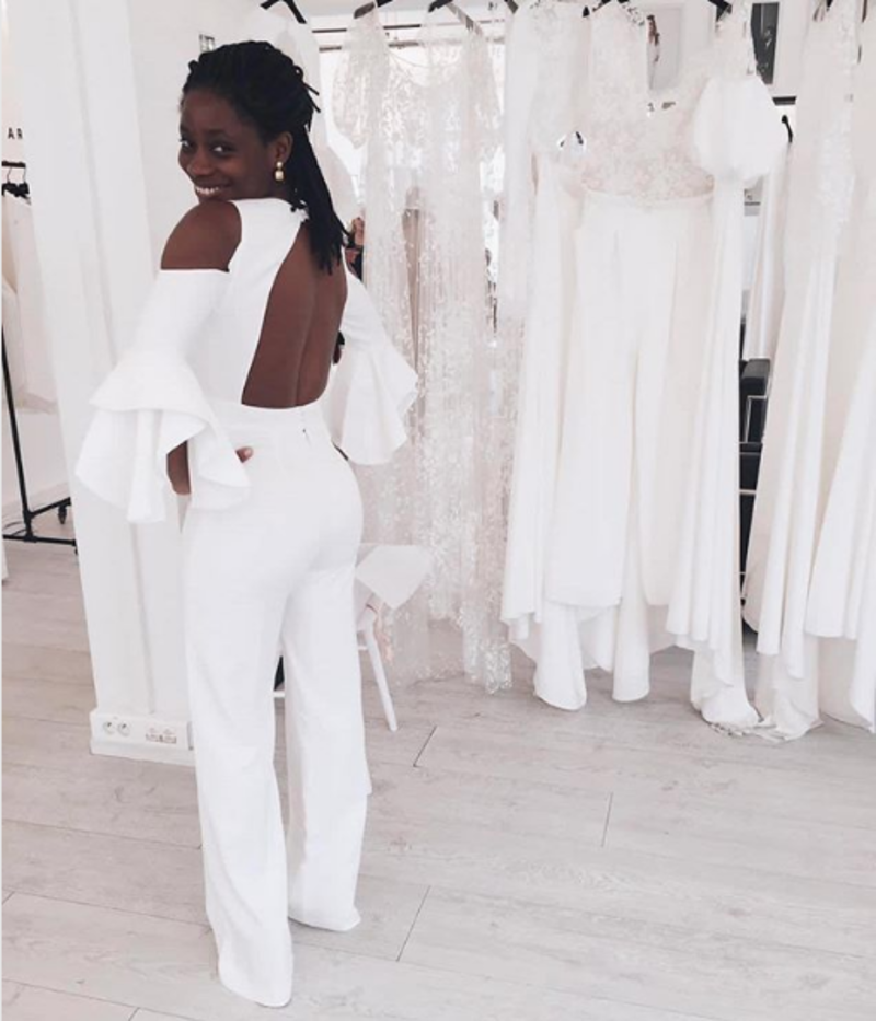 Mulher experimentando roupas em uma loja de vestido de noiva. Ela veste um macacão branco, frente única, e com manga longa e um detalhe solto nas pontas. Ela sorri para a foto.