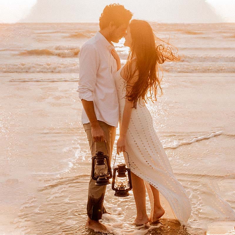 Casal com look pré-wedding segurando lanternas na praia com os pés na água