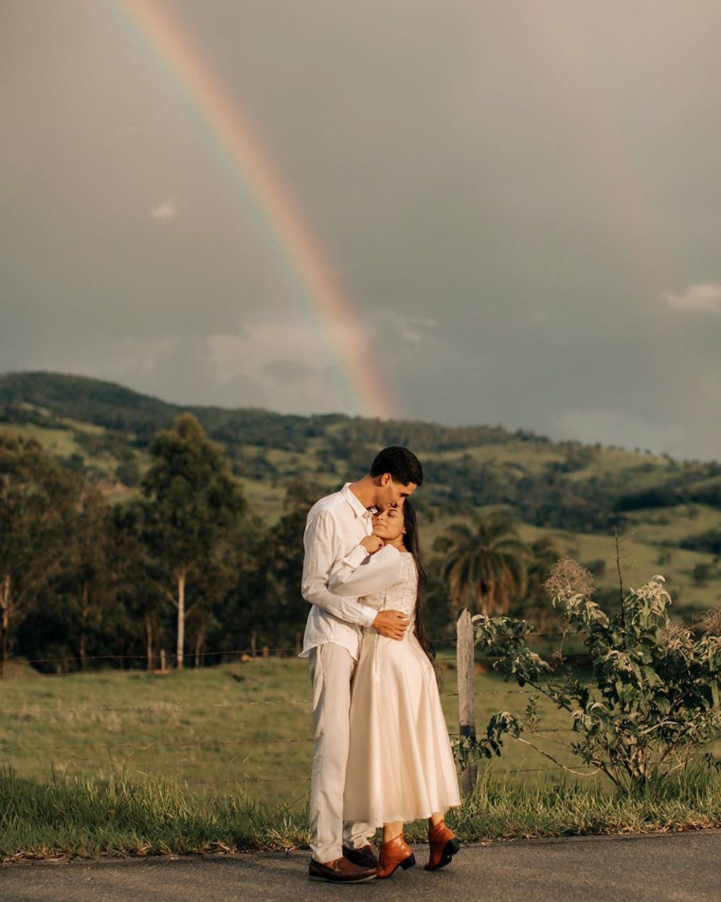 Casal em pose romântica com arco-íris ao fundo, ambos de branco, ilustrando um possível look pré-wedding