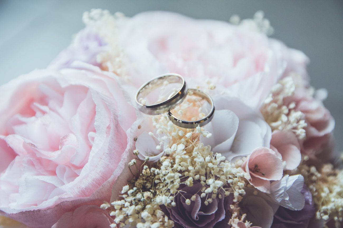 Um par de alianças em cima de um buquê de flores brancas e rosas. Imagem disponível em Unsplash.