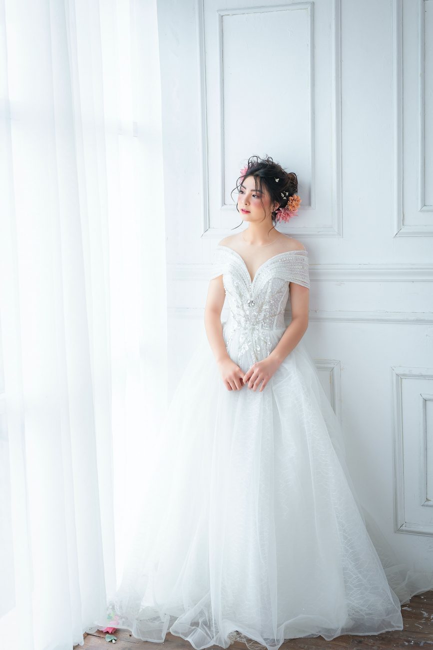 Noiva, vestida de branco, posa para foto em um fundo branco, ao lado de uma cortina. Imagem disponível em Pexels.