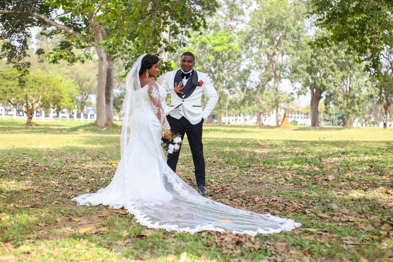 Homem e mulher vestidos de noivos posam para foto em um parque com gramado verde. Imagem disponível e Pexels.