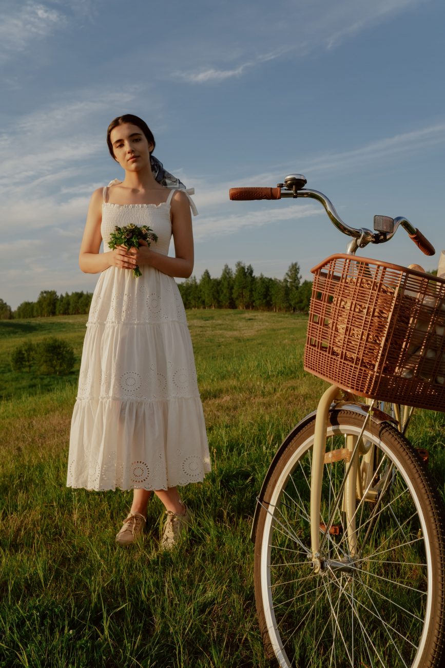 Mulher com vestido branco, segurando um buquê, ao lado de uma bicicleta em um campo verde.