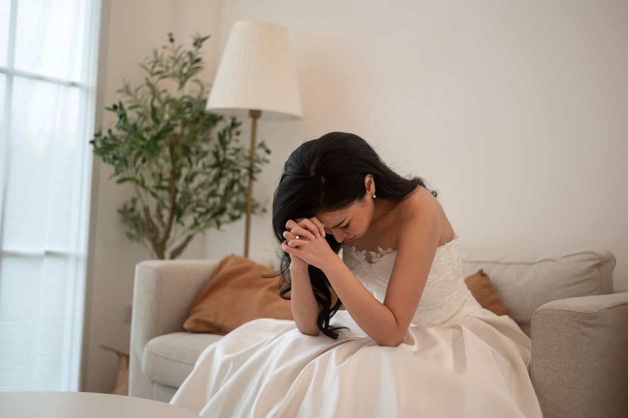 mulher vestida de noiva, sentada em sofá branco, com mãos em oração e cabeça baixa