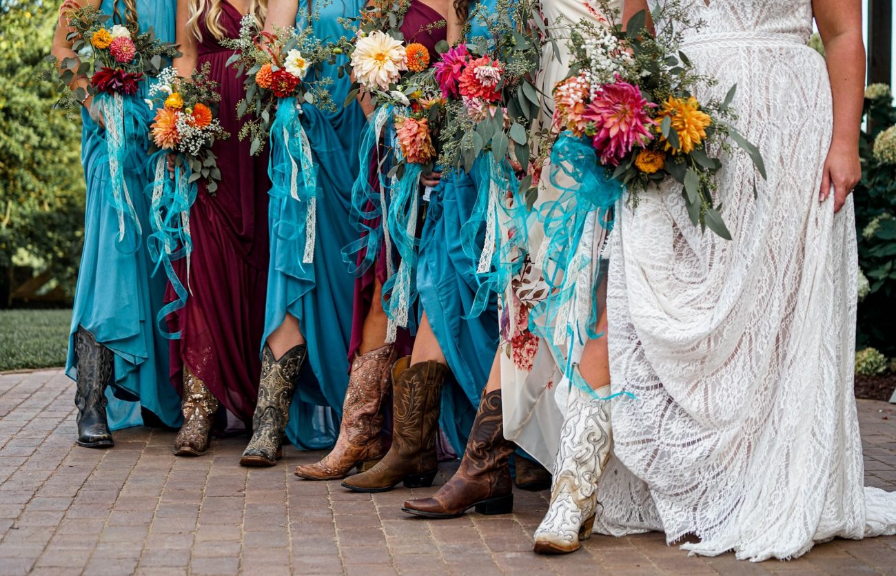 Casamento Temático Musical: noiva vestida de branco e madrinhas de azul e vinho, segurando flores coloridas e usando botas de cowboy