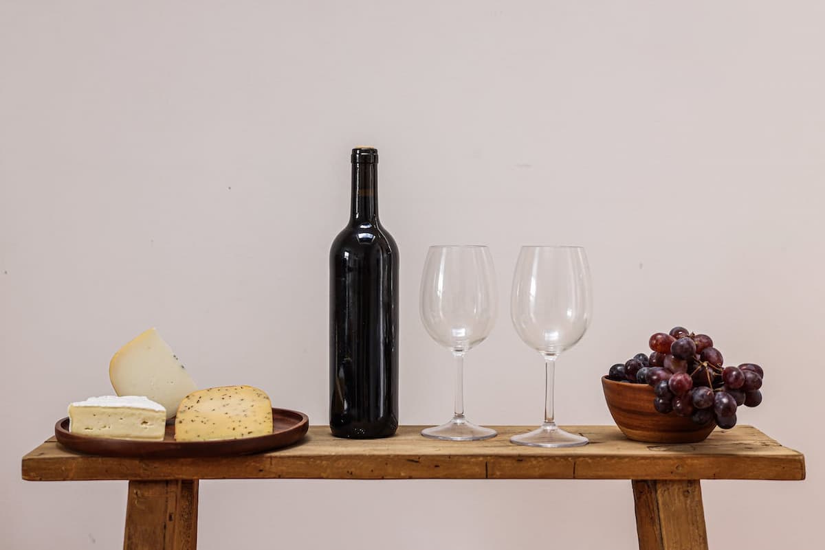 Imagem de um vinho, taças e queijos em uma mesa de madeira
