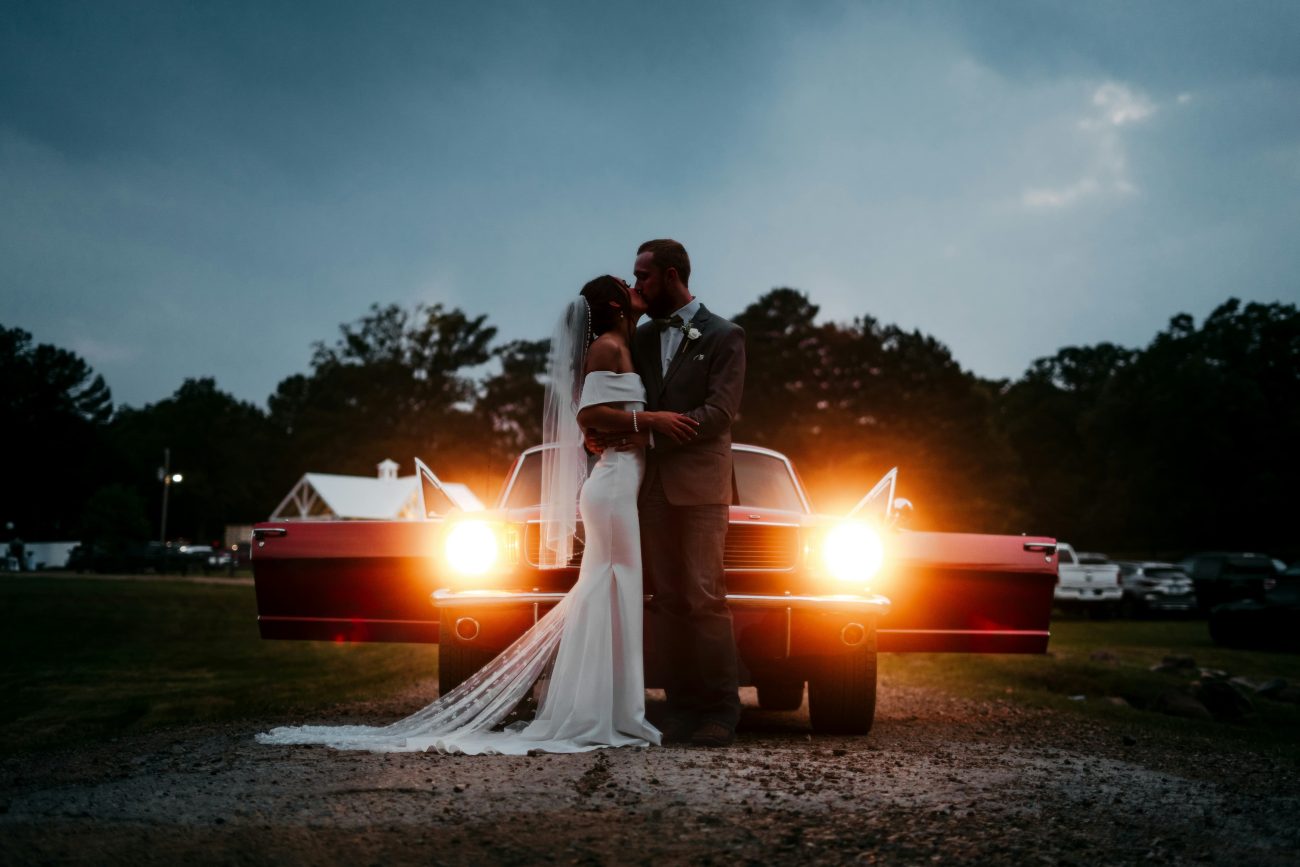 casal de noivos se beijando na frente de carro antigo com faróis acesos