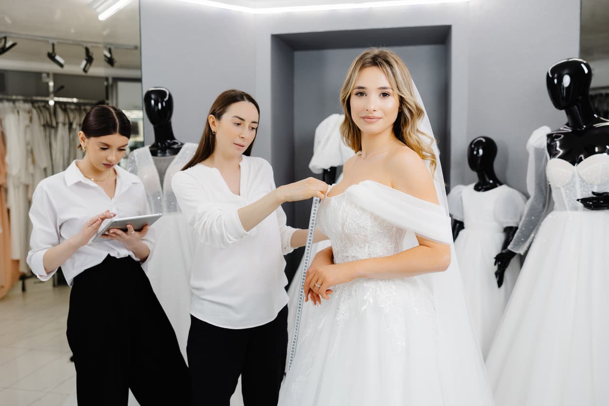Noiva em ateliê, com uma costureira conferindo as medidas do seu vestido de casamento.