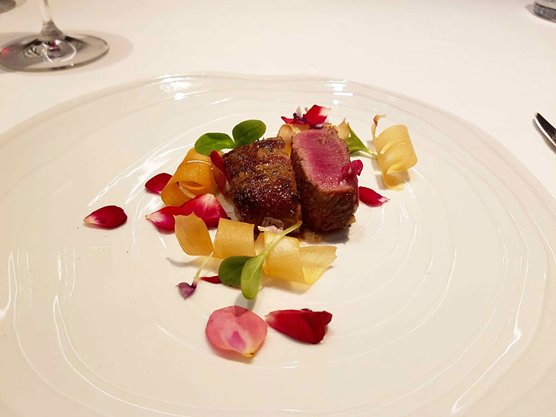 Prato branco com carne vermelha assada, com pétalas de flores ao redor.