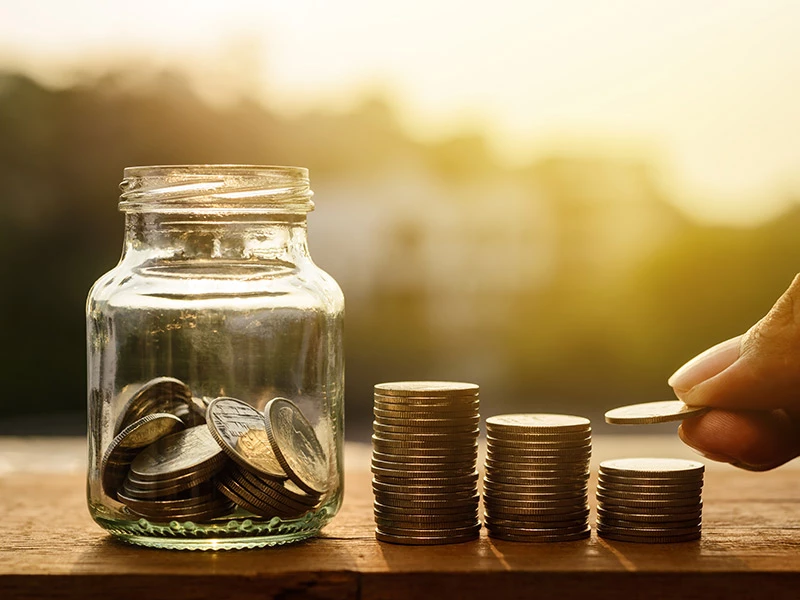 Pote de vidro com moedas dentro e pilhas de moedas ao lado.