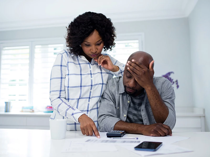 Casal negro, homem sentado e mulher em pé com cotovelo apoiado no ombro do homem. Ambos observando uma tabela impressa em cima da mesa.