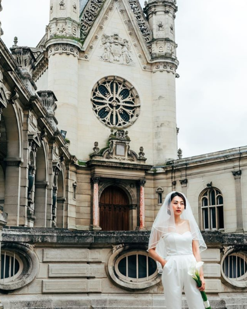 noiva de macacão na frente da igreja, com véu longo e flores na mão