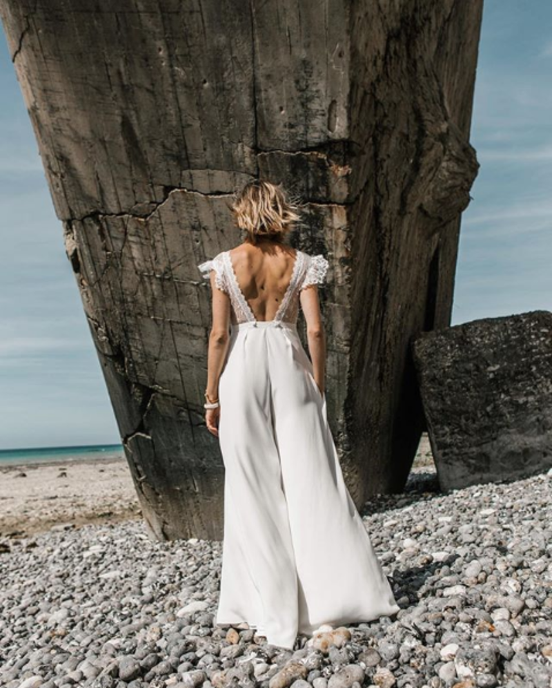 Noiva de macacão na praia de pedras, olhando para o cenário, de costas para a câmera. O macacão tem o detalhe das costas abertas