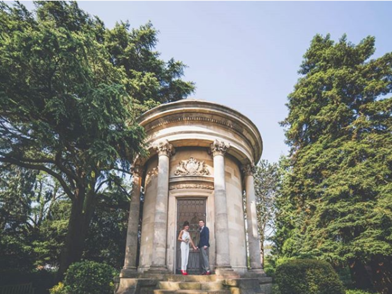 Casal se casando ao ar livre, em uma área cercada de árvores e verde, ele vestindo o terno e ela um macacão branco. 