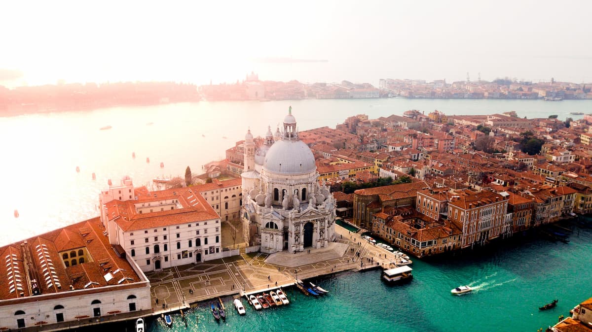 Foto aérea de Veneza mostrando um grande edíficio e o grande canal|Pessoas andando durante o dia em um grande espaço em Veneza|Imagem de uma mesa de casamento com decoração de flores e copos e pratos|Imagem de barcos no grande canal de Veneza no pôr do sol|Pessoas andando de barco no grande canal durante o dia