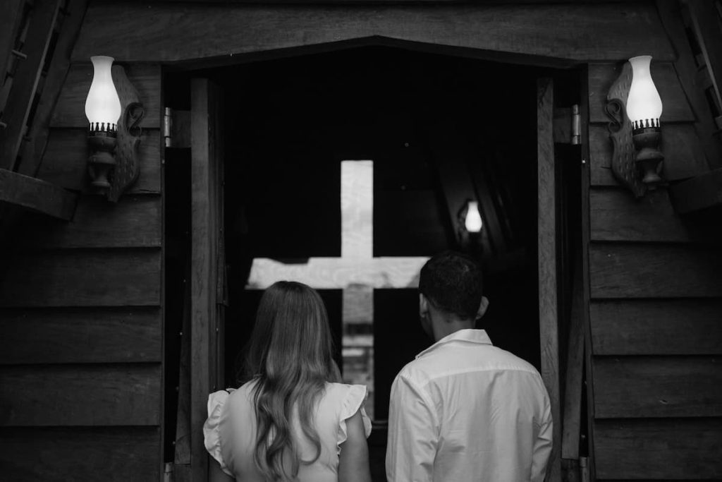 Foto em preto e branco de casal de noivos em igreja com cruz.|||