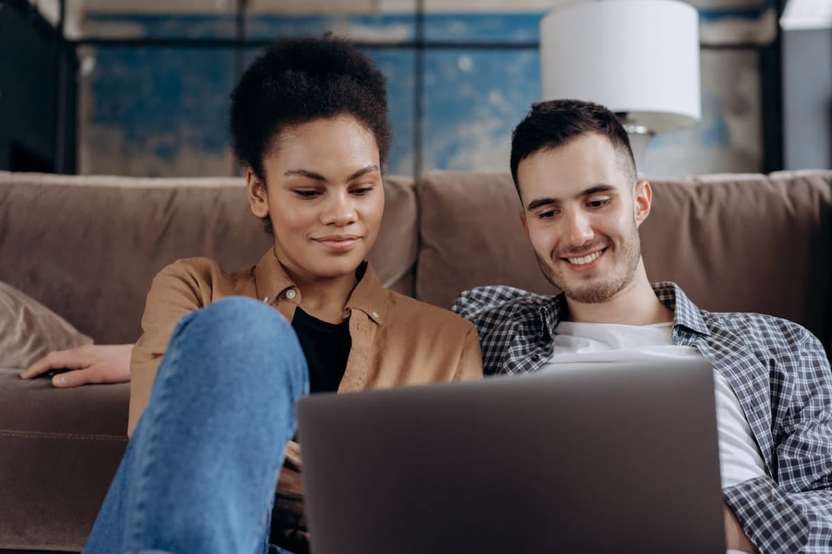 Imagem de um casal olhando para o computador sentados no sofá|Imagem de um papel com a lista de presentes de casamento do lado de um computador na mesa