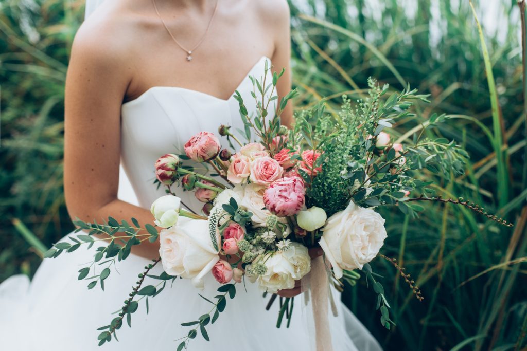 noiva com vestido branco e buque de flores coloridas na mão|flores rosas e brancas com alianças de casamento em cima|casal de noivos em frente a parede de pétalas rosas