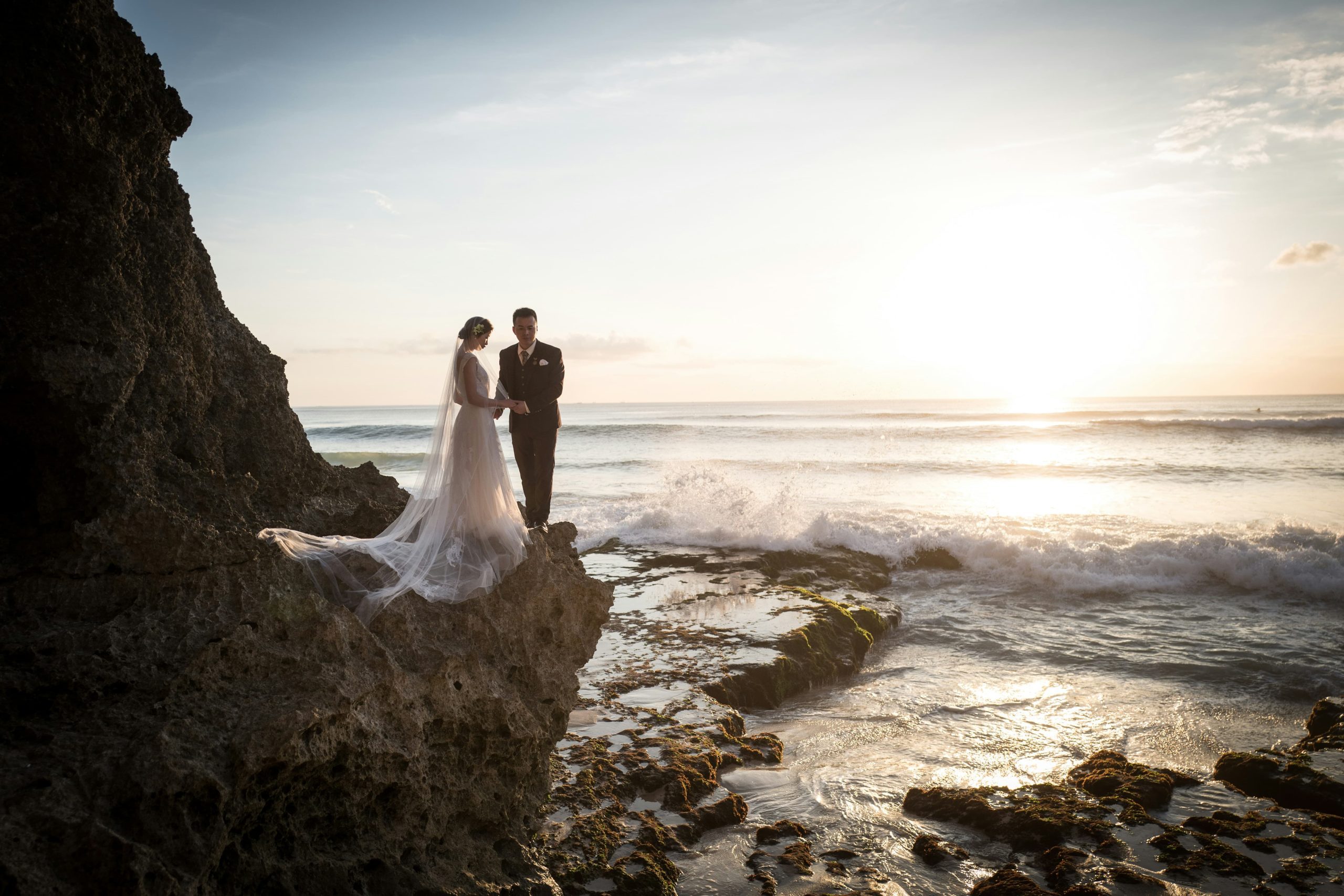 Casal de noivos em pedra na beira do mar|recepção de casamento na areia. Foto mostra areia branca com cadeiras brancas organizadas em fikeiras e decoradas com flores rosas. Mar azul ao fundo|