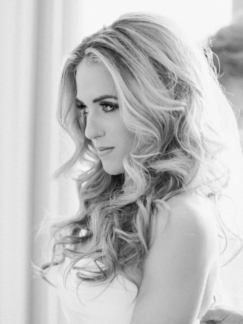 Penteado para noiva solto com mechas torcidas e um pequeno arranjo floral, ideal para casamentos no campo