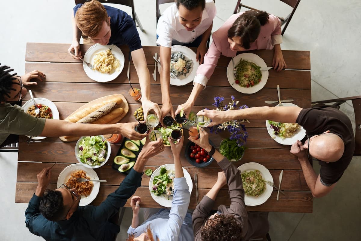 Amigos brindando em uma mesa com diversas comidas||Amigos conversando ao redor de uma mesa com comidas|Imagem de uma bancada com bebidas e comida e pessoas ao redor conversando