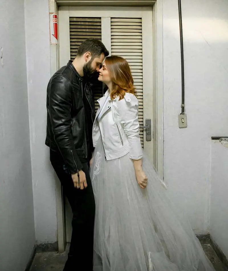 Casal em ensaio pré-wedding: ela de vestido branco longo com jaqueta de couro, ele de look preto com jaqueta