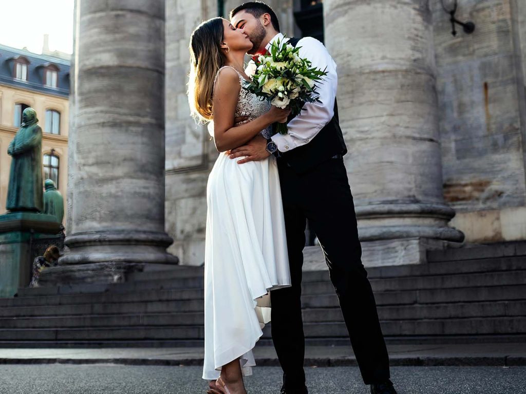 Casal com vestido de casamento e buquê de flores se beijando.